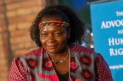 [ai] A woman with curly hair wearing a colorful patterned blouse and a decorative headband, looking directly at the camera with a neutral expression, in front of a blue banner with text related to human rights advocacy.